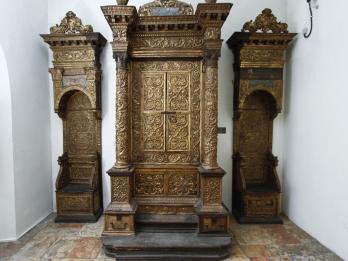 Wooden Torah ark decorated with carvings, columns, and capitals, flanked by two similarly decorated chairs. 