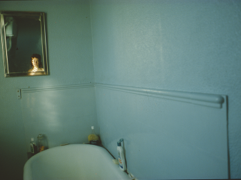 Photograph of bathroom wall with bathtub and small mirror above that reflects a face. 