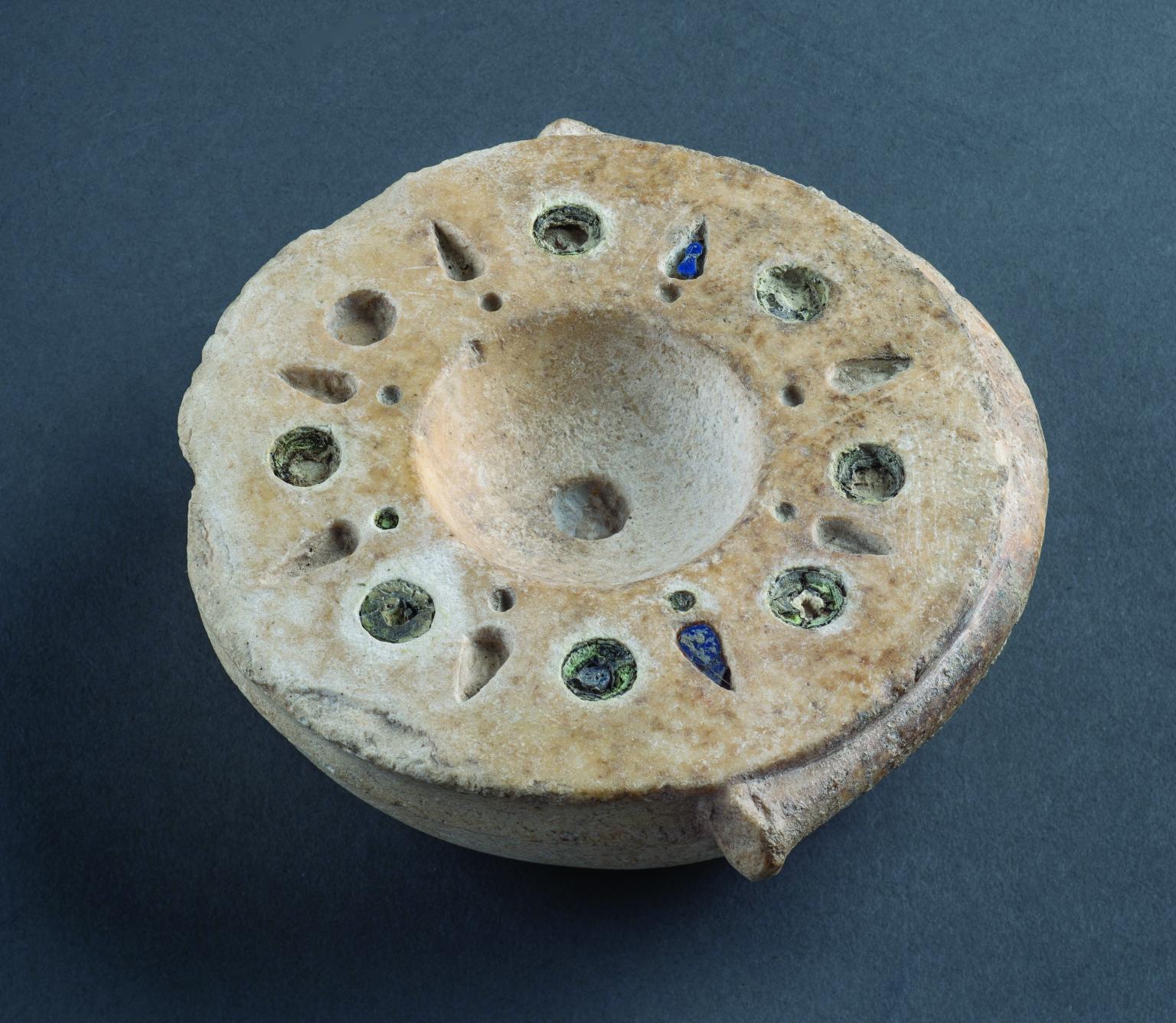 Limestone bowl with mosaic decoration around rim with alternating circular and bud-shaped inlaid blue and brown glass. 
