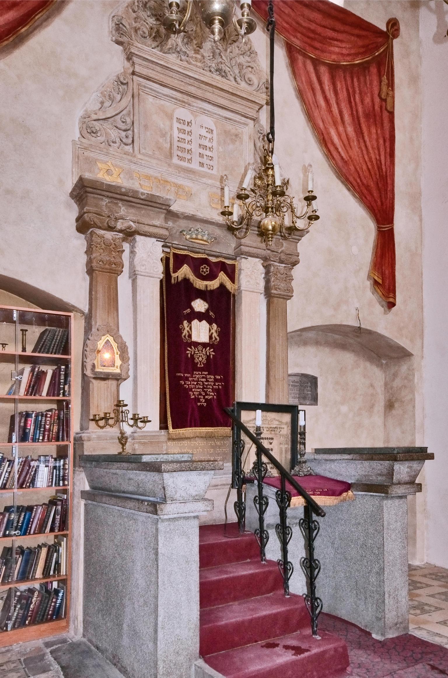 Photograph of Torah ark built into stone wall and covered with cloth, with small staircase in front and bookshelf to the left.