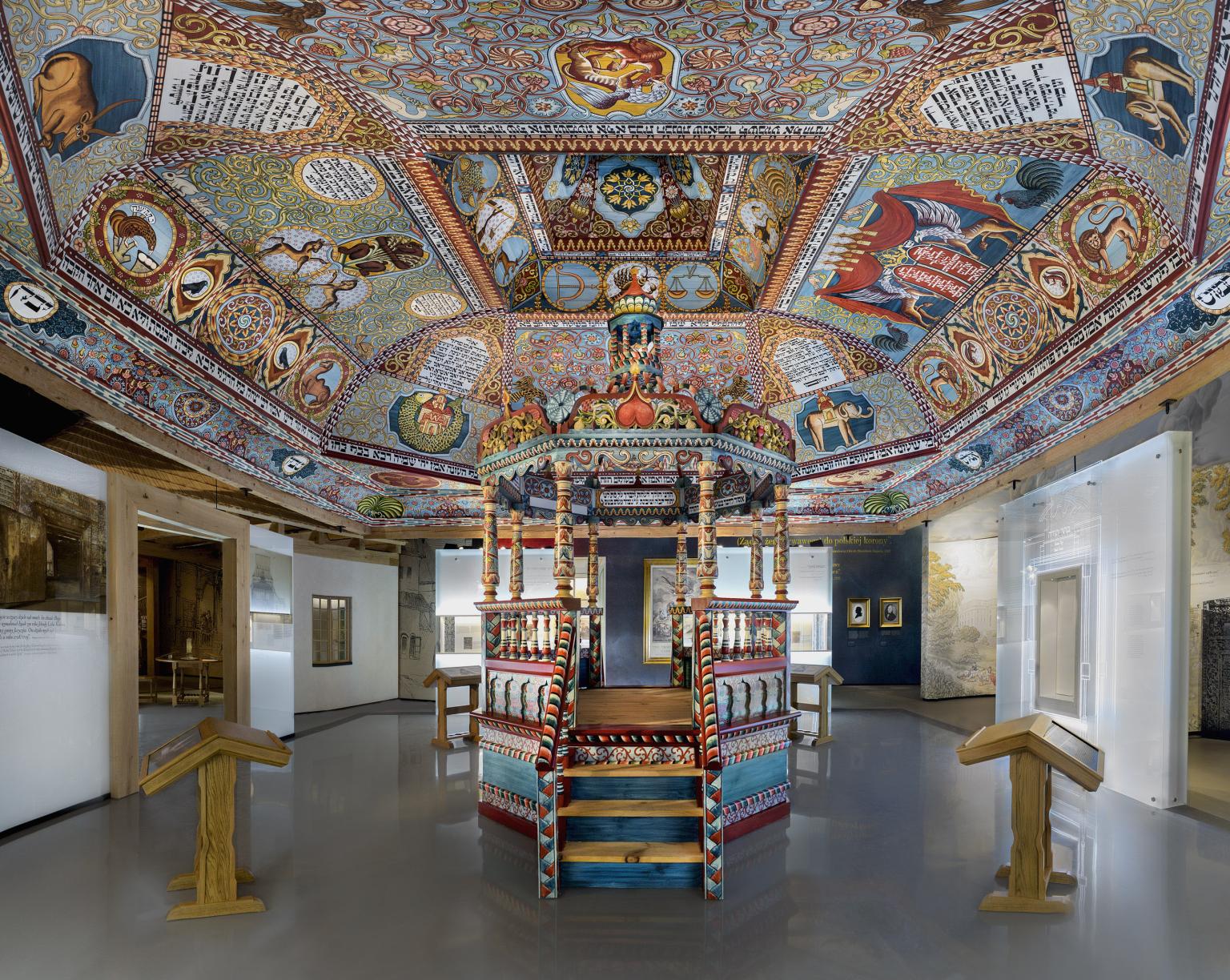Photograph of room with mural ceiling of floral patterns, animals, and panels of Hebrew text, and painted canopied platform in center of room.  