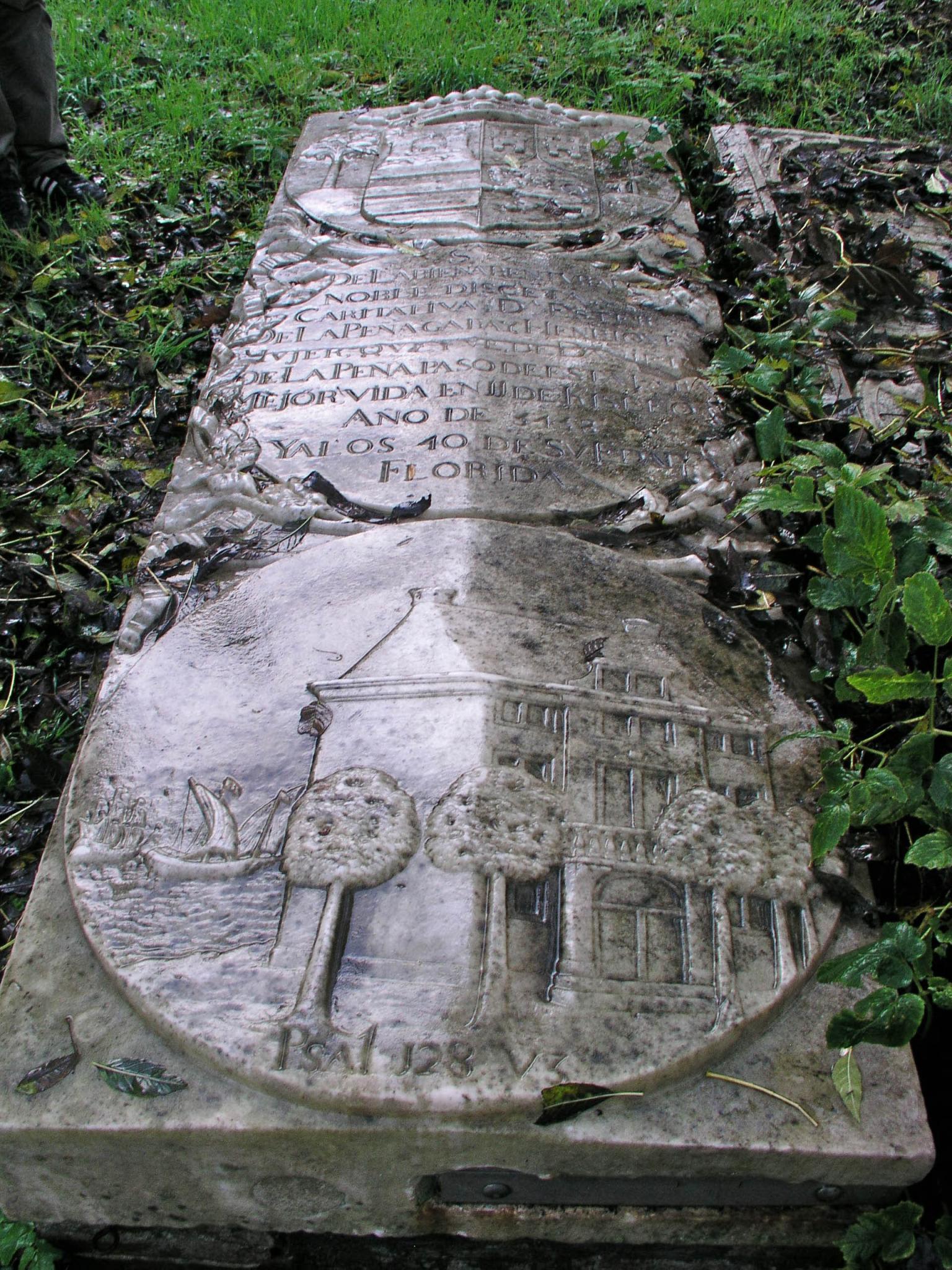 Tombstone flat on ground depicting coat of arms above Spanish inscription and relief of house surrounded by trees and next to sailboat.