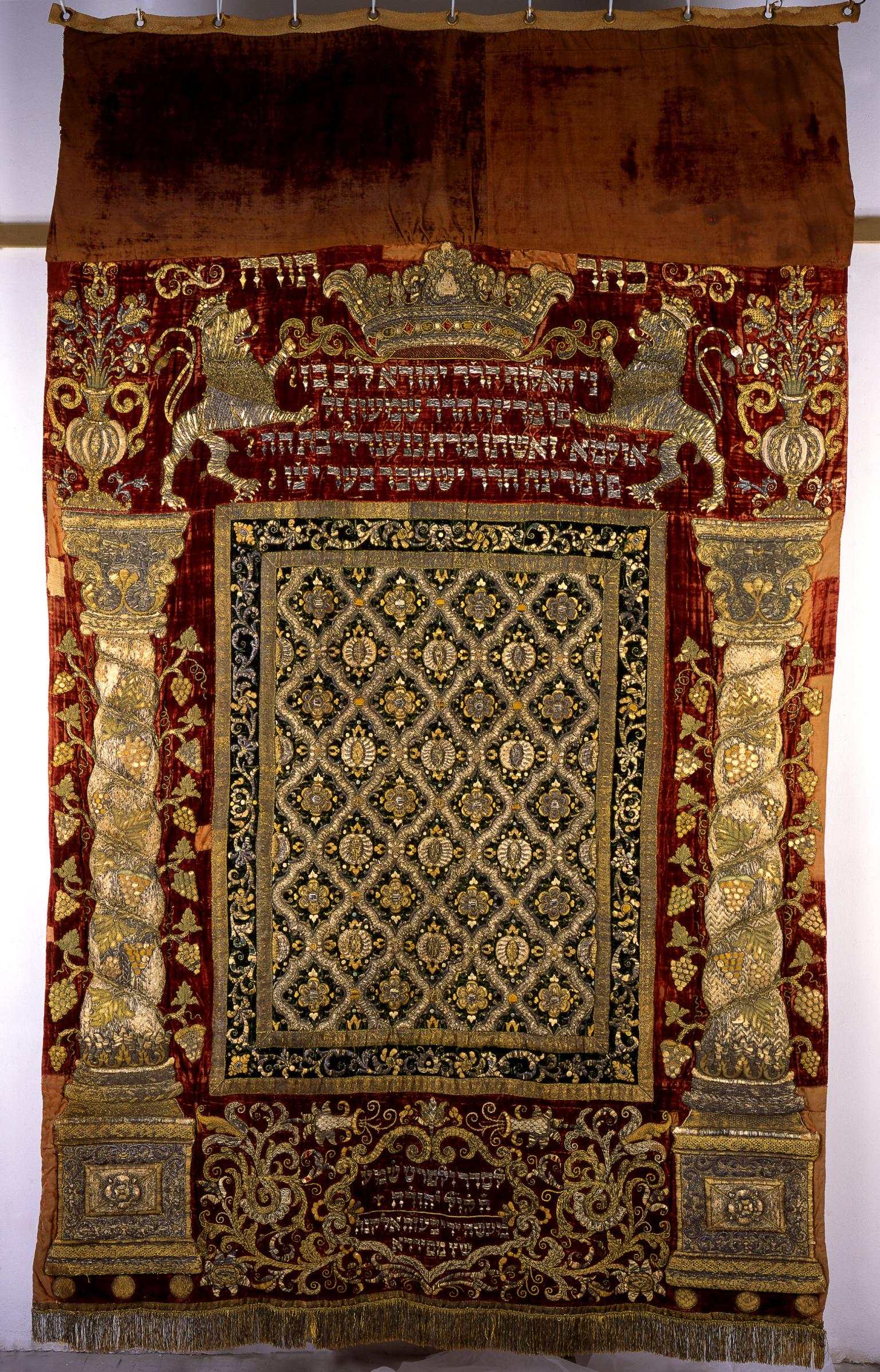Cloth with embroidered columns on either side, pattern in the middle, and Hebrew text between two lions and crown on top.
