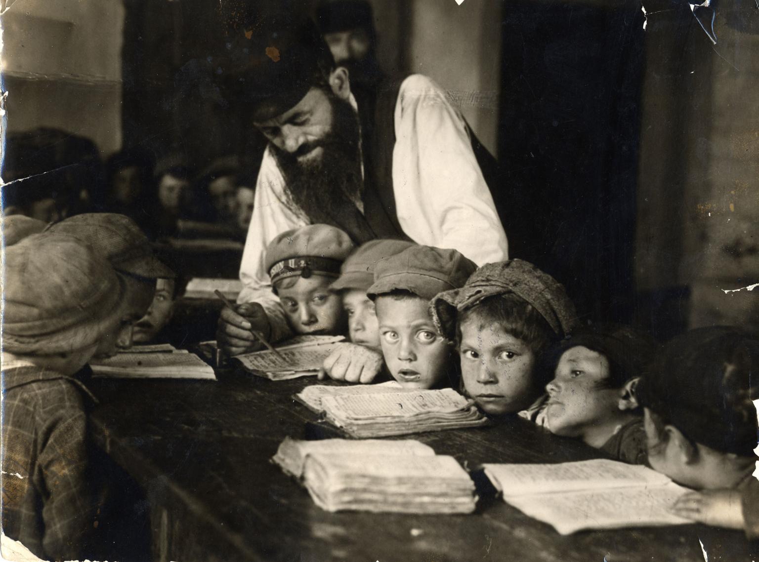 Photograph of young boys sitting at a long table with books open as teacher leans over them to teach.