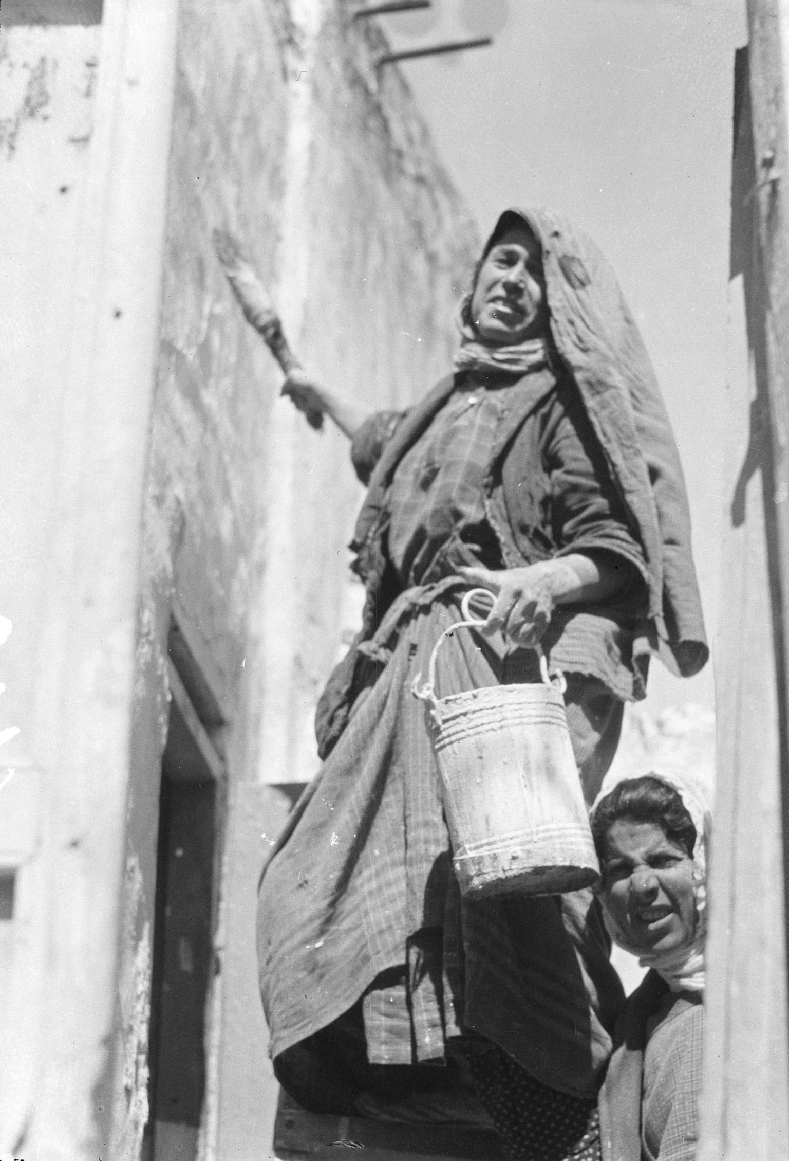 Photograph of woman in headscarf looking toward camera, holding bucket and paintbrush next to wall, with woman behind her looking at camera.