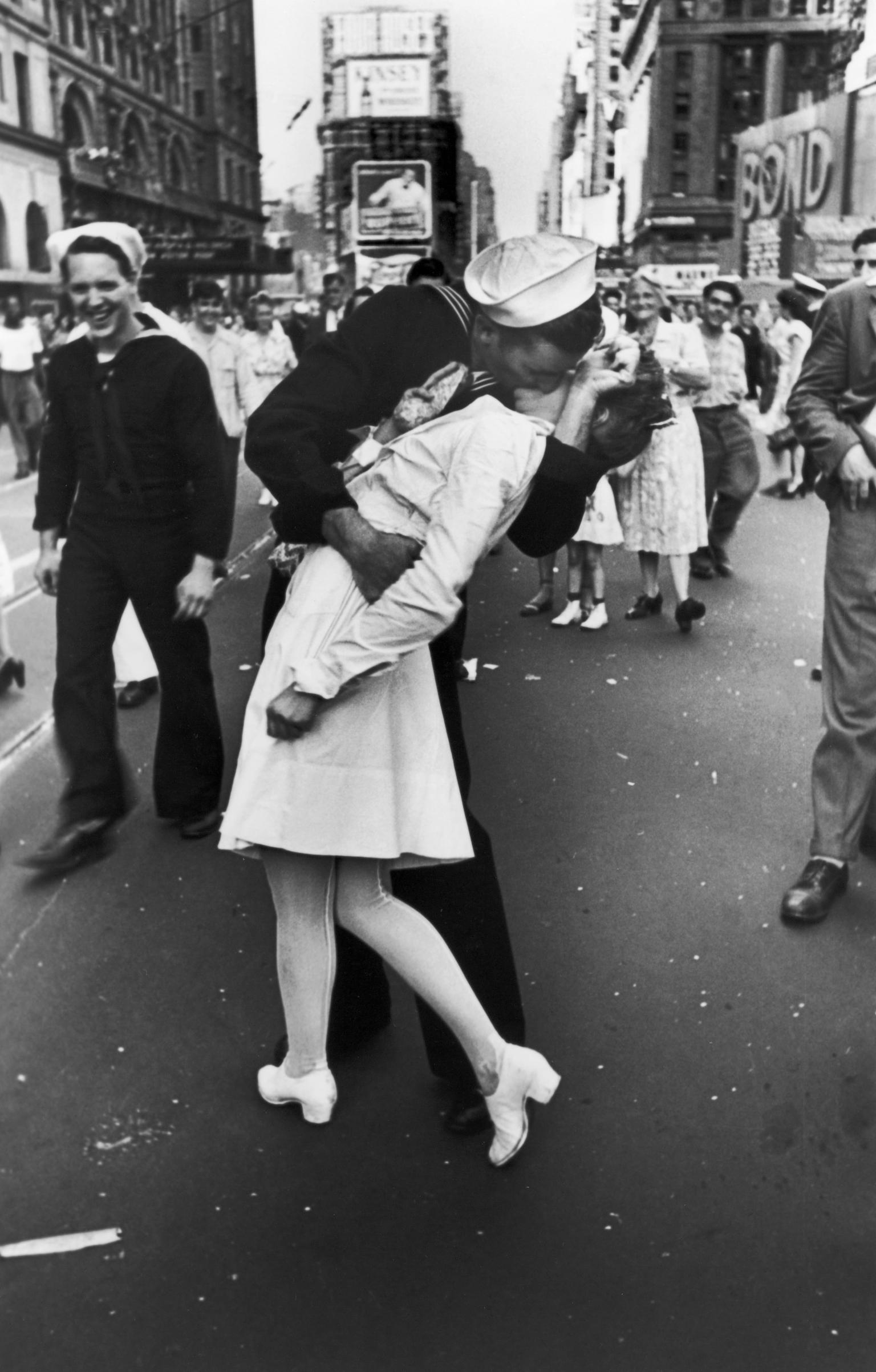 Photograph of sailor grabbing and kissing a woman in a busy city street with people and buildings in background. 