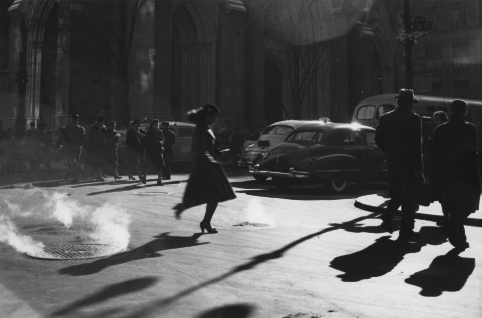 Photograph of woman in a dress and hat walking across a city street with cars, people, and buildings in the background.