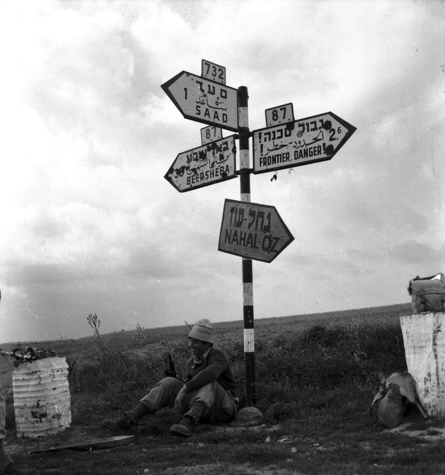 Outdoor photograph of signpost with signs pointing different directions with town names and information in Hebrew, English, and Arabic, and smiling man holding a rifle sitting on the ground beneath the sign.