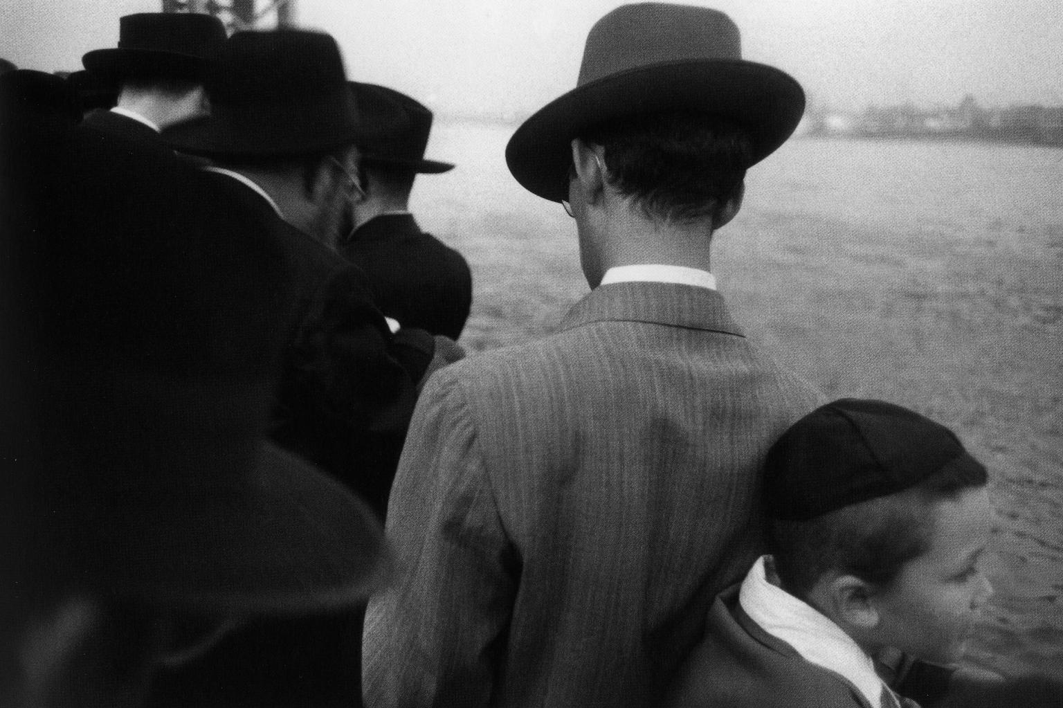 Photograph of men in hats and suits and one boy in skullcap outside with their backs turned away from the camera, looking toward the river.