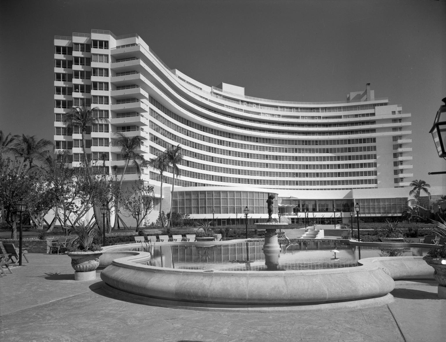 Photograph of curved exterior of a hotel with pool and fountain in the middle.