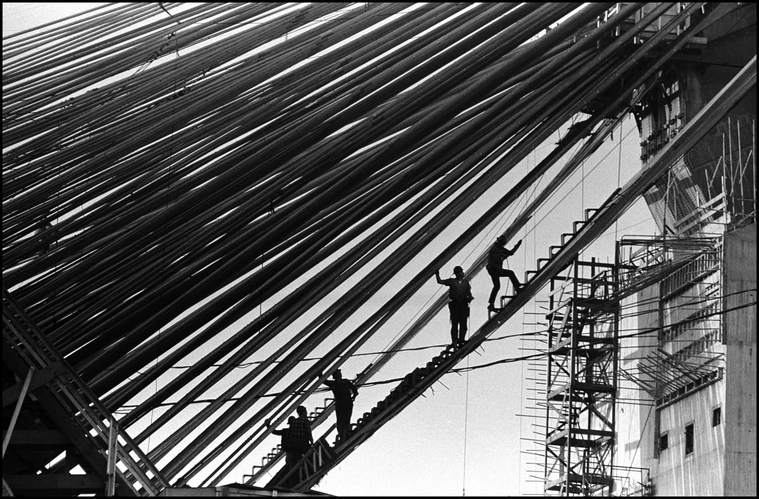 Photograph of men silhouetted on a steep ladder that bisects the photograph on a diagonal, below cables that run across the photograph, and tall building in background.
