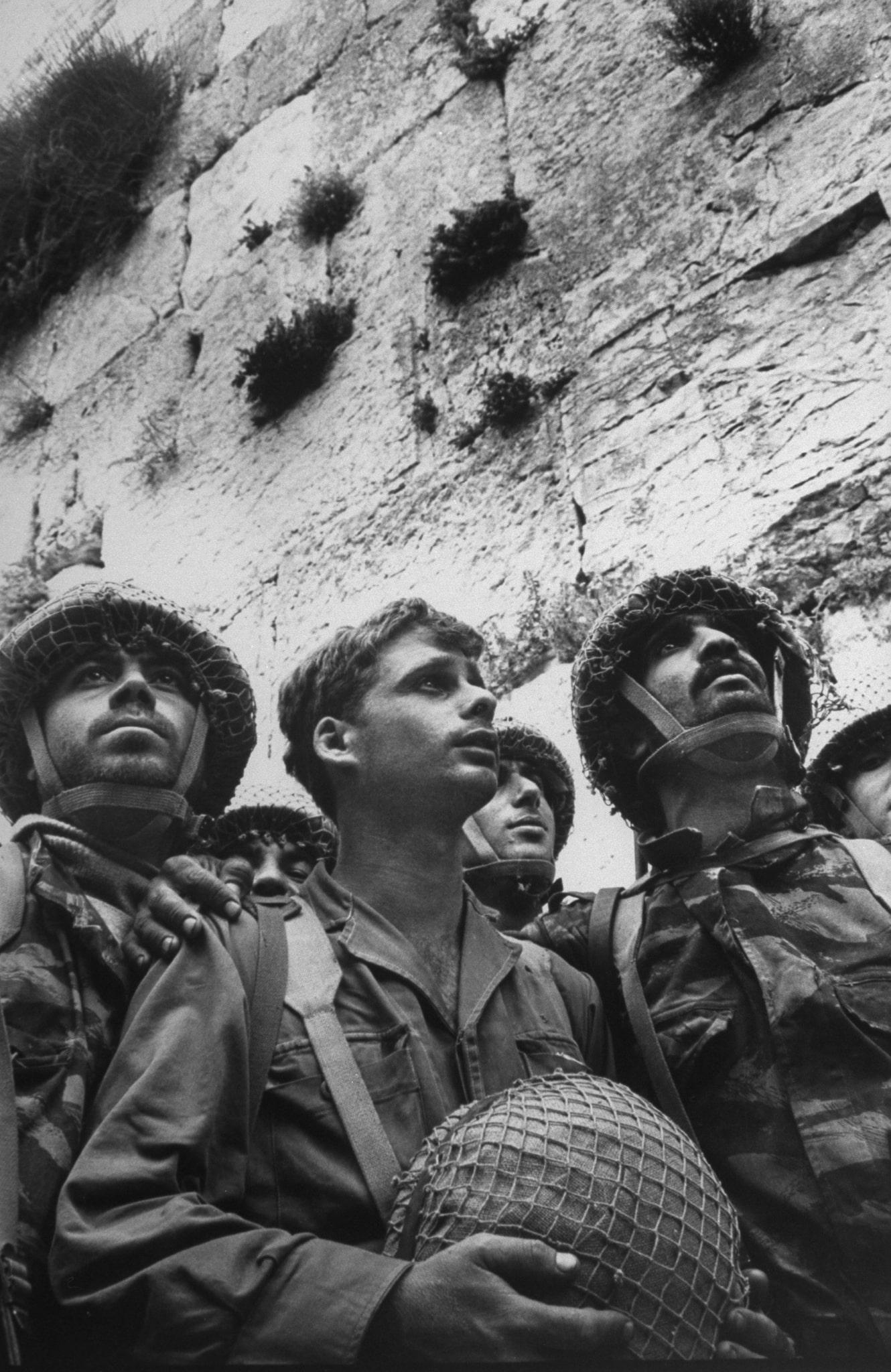 Photograph of three soldiers in uniform looking to their left standing in front of the Western Wall, with central soldier holding helmet in his hands.