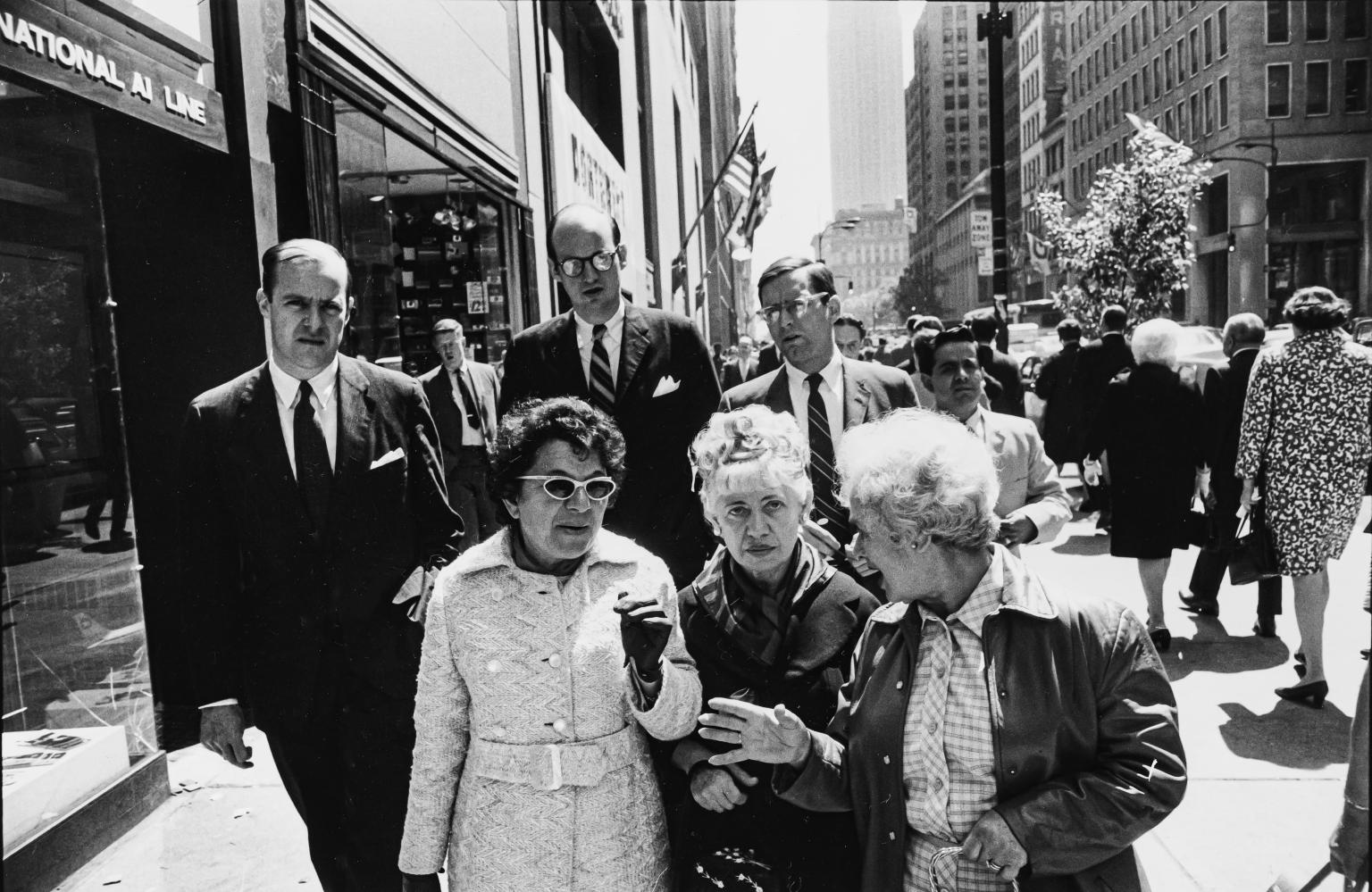 Photograph of three elderly women walking and talking on busy city sidewalk with men in suits behind them.