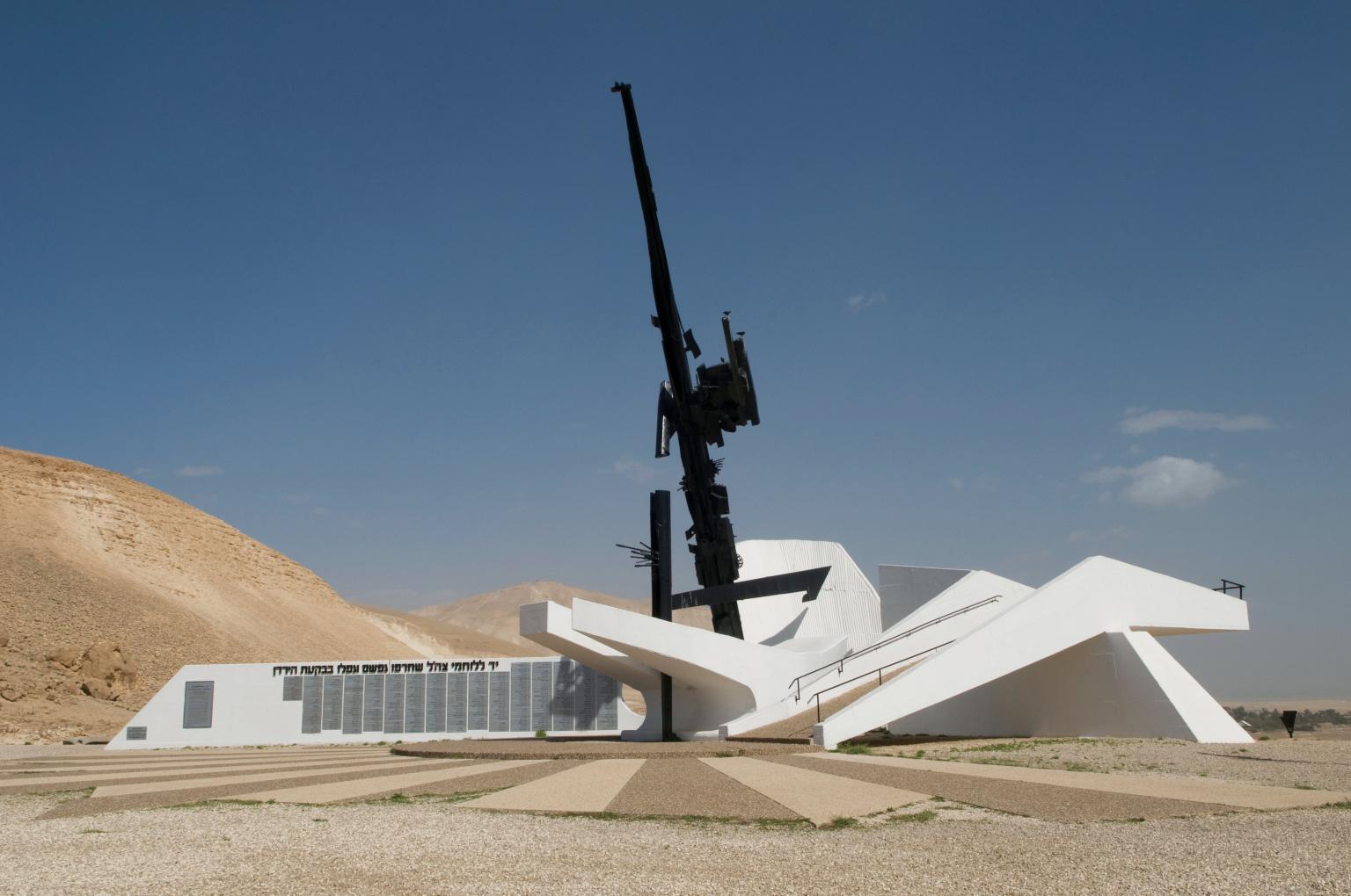 Cement and steel monument resembling a large firearm in a white base next to a building, surrounded by sand dunes.