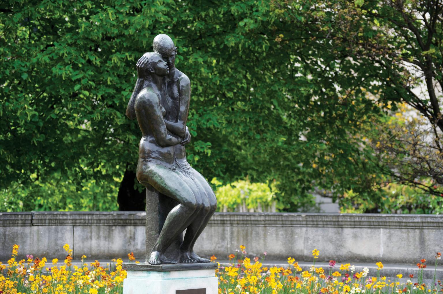 Bronze sculpture of young couple, seated side-by-side and cheek-to-cheek in a garden. The ground is covered by leaves and there are many trees in the background beyond a low wall.