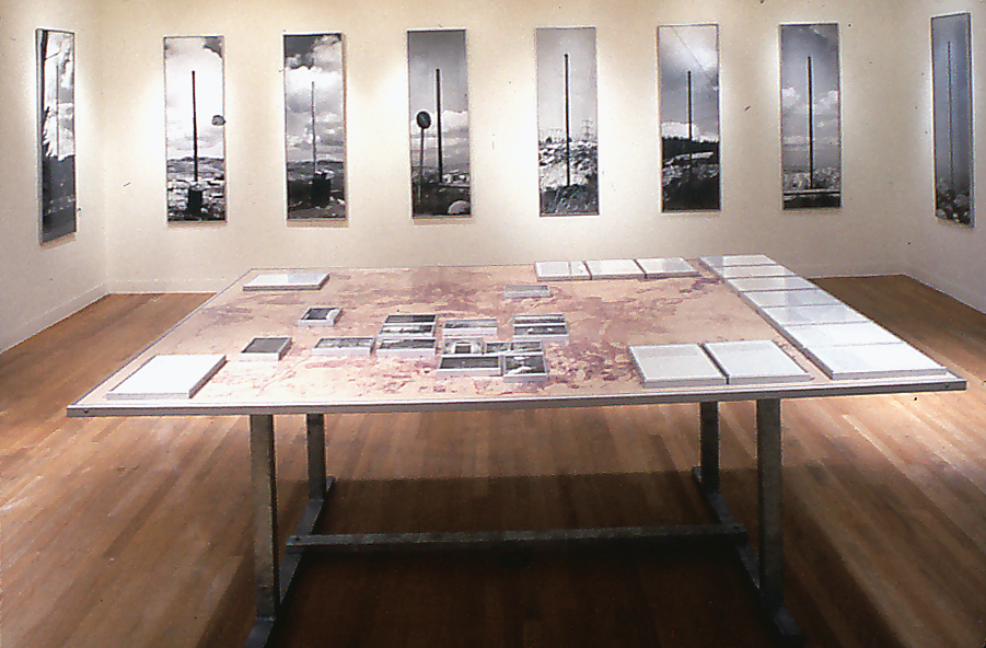Installation showing set of eight photographs arranged on back wall, and table in foreground with fourteen photographs and fourteen texts lying flat.  