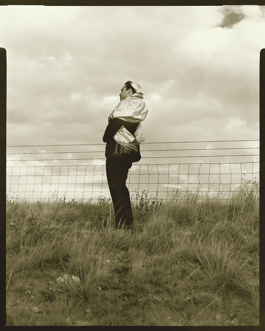 Full body photograph depicting man in kippah and prayer shawl standing in a field next to fence, facing to the side.