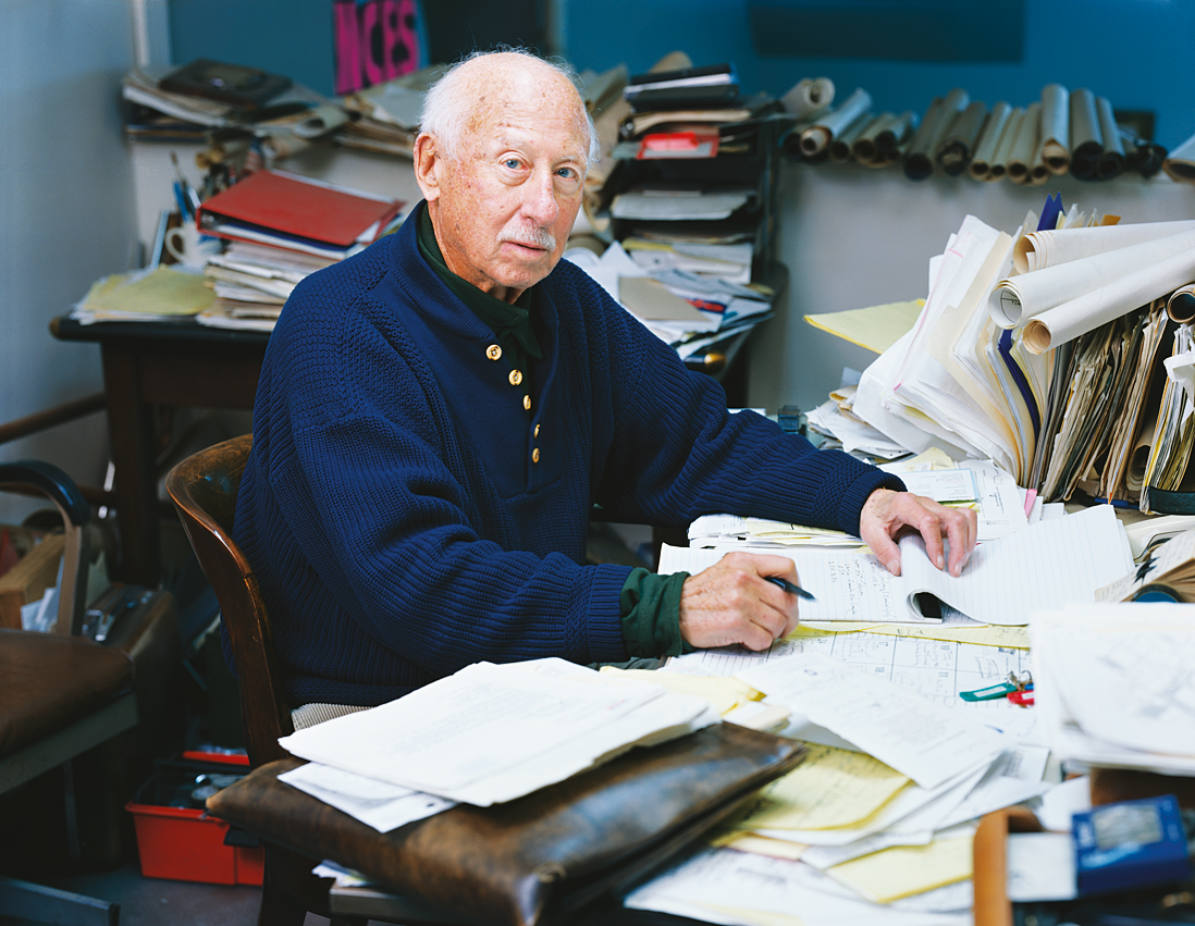 Photograph featuring older man facing viewer and seated at cluttered desk.