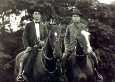 Two men wearing jackets and hate are seated on horses.