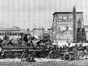 Enslaved people pull a cart bearing a sphynx past monumental Egyptian buildings decorated in heiroglyphs.