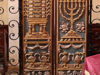 Set of curved doors with carving of candelabrum, peacock-shaped vessels, and basket of fruit and flowers on right side and table with compartments, peacock-shaped vessels, and basket of fruit and flowers on left side. 