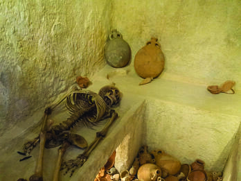 Interior room with ledges, one of which holds a skeleton, and a floor covered with pottery.