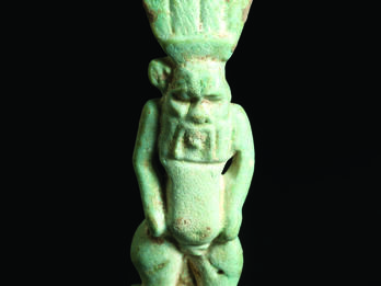 Frontal and back faience amulet of figure who is nude with feathered headdress and a grotesque face and beard.
