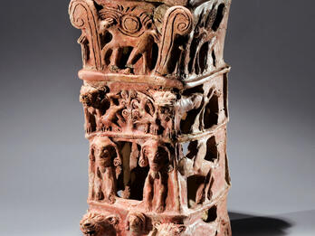Ceramic offering stand with flat top and four layers of decoration: bottom layer of nude female with raised arms flanked by lions, second layer of two winged figures, third layer of goats eating leaves off tree, and fourth layer of four-legged animal with winged sun disk and palm fronds. 
