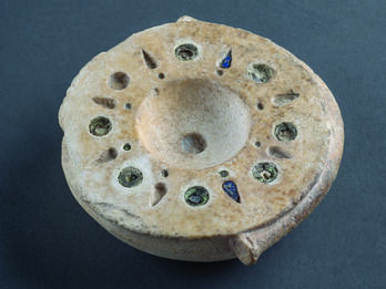 Limestone bowl with mosaic decoration around rim with alternating circular and bud-shaped inlaid blue and brown glass. 