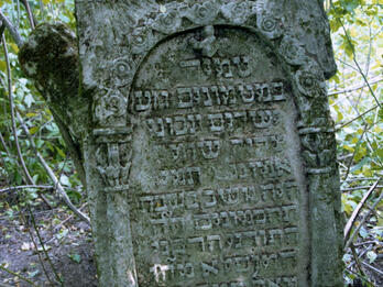 Tombstone with Hebrew inscription engraved with columns, flowers in curved shape above text, and wings at the top of the stone. 