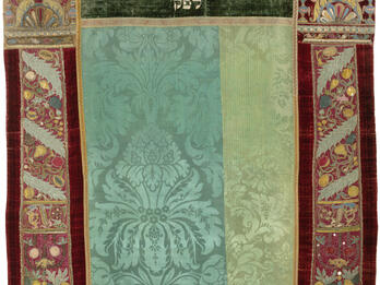 Embroidered curtain of columns with crowns on left and right sides and decorative floral motifs throughout, and Hebrew text in top center.