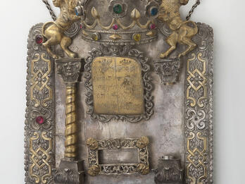 Shield with decorative border inlaid with semi-precious stones and adorned with lions and crown at top, column on left and plaque in middle with Hebrew text. 