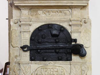 Carved wall unit with decorative borders and crowns, locked metal box in middle, and Hebrew text underneath. 