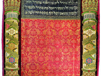 Embroidered curtain of columns with crowns on left and right sides and decorative floral motifs throughout, and Hebrew text in top center.