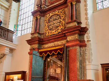 Photograph of tall ark holding two Torahs built into interior wall with Hebrew writing and surrounded by short fence with Stars of David. 