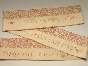 Torah Binder Rectangular cloth, folded in two places, with Hebrew text embroidered on bottom half of cloth and floral border on top half.