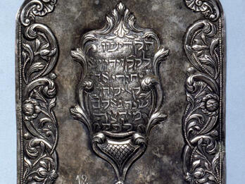Silver shield of decorative border with Hebrew text in center.