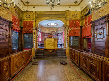 Photograph of room lined with narrow wooden pews and set of stairs at far end of room leading to gilded podium.