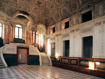 Interior of room with columns and short double staircase at one end and pews along wall.