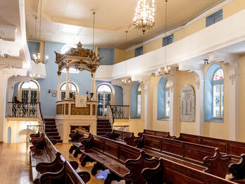 Photograph of room with high ceiling, tall podium up short staircase with golden canopy, and rows of wooden benches. 