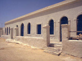 Exterior photograph of stone building with arched windows, wraparound patio with stone columns, and dirt ground.