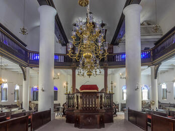 Photograph of room with central podium, two-story columns, benches facing podium, and balcony above, with sand floor. 