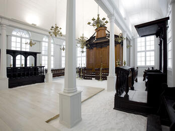 Photograph of rectangular room with arched windows, floor covered in sand, wooden cabinet on far wall, wooden benches on left side and along walls, and wooden lectern on right side. 