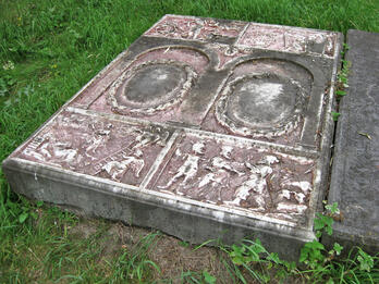 Tombstone flat on ground of panels of scenes with figures and animals above and below arch-enclosed Hebrew inscription.