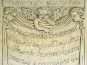 Tombstone of three cherubs and central Portuguese inscription, with engraving of wilted flower, skull, hourglass, and sun, with top panel of engraving of man riding in a procession through street and Hebrew text across the middle. 