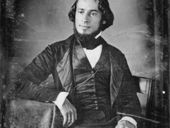 Portrait of man in suit facing slightly angled, seated with his arm resting on the table.