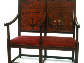 Two-seater bench with central armrest, decorated on the seat backs with ritual objects and Hebrew text.