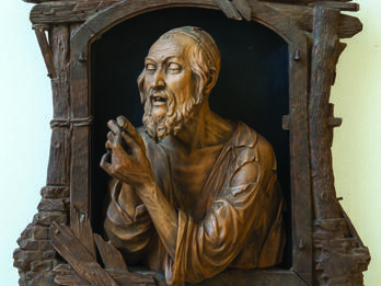 Wood-carving of a man in skullcap examining an object in his hands.