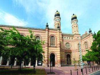 Photograph of exterior facade of building with minarets, circular windows, and decoration, and trees in courtyard. 