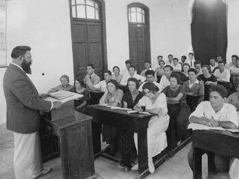 Photograph of classroom with teacher at podium and men and women at rows of desks.